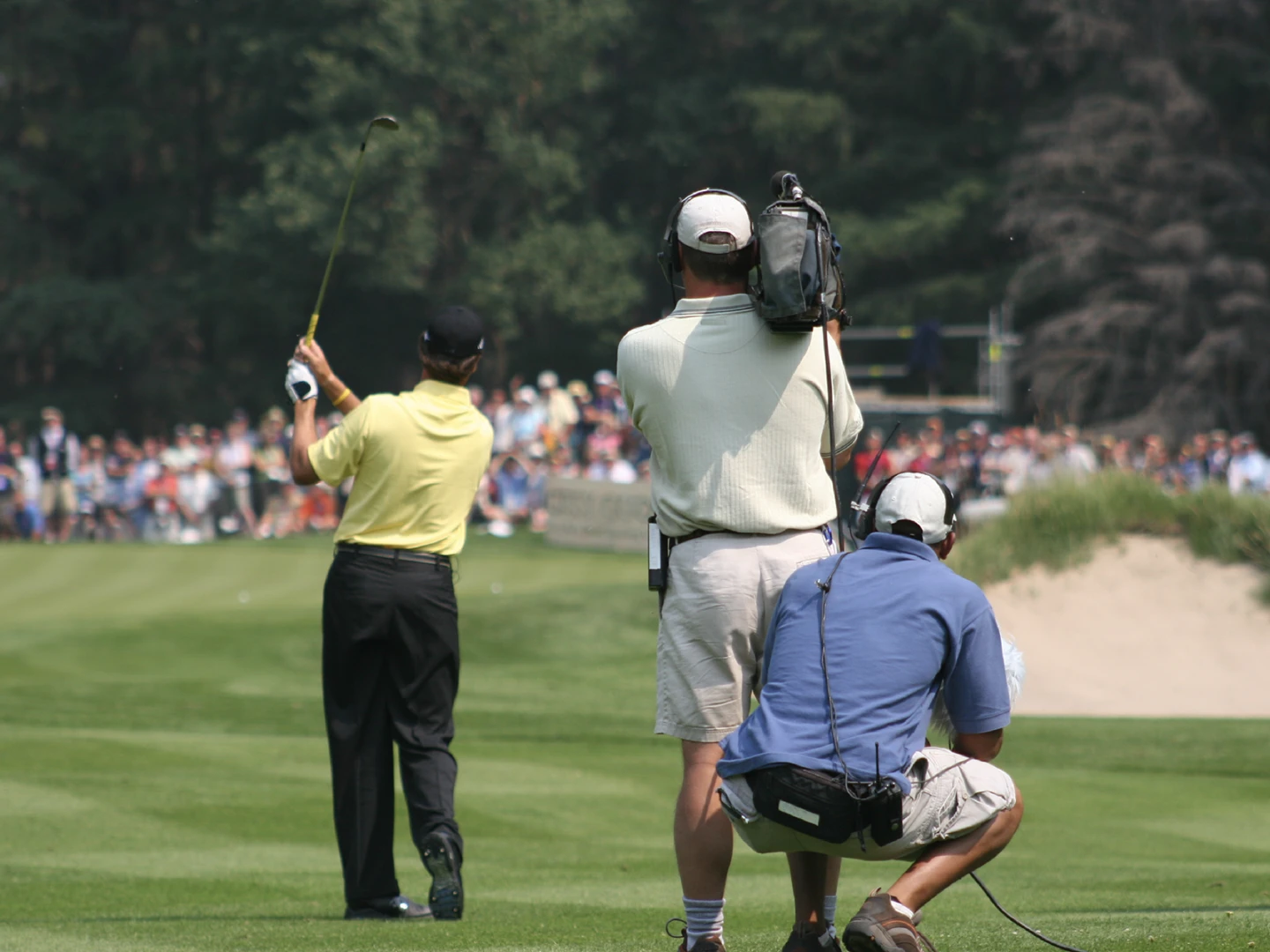 image of professional golfer playing approach shot on golf course with camera crew behind and spectators overlooking green